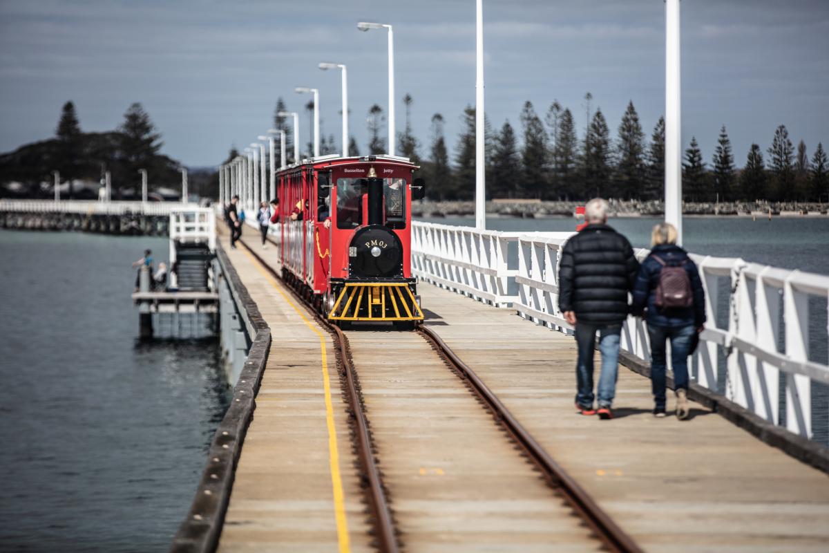 Red train driving along Busselton Jetty with people Walking along the jetty nearby. 